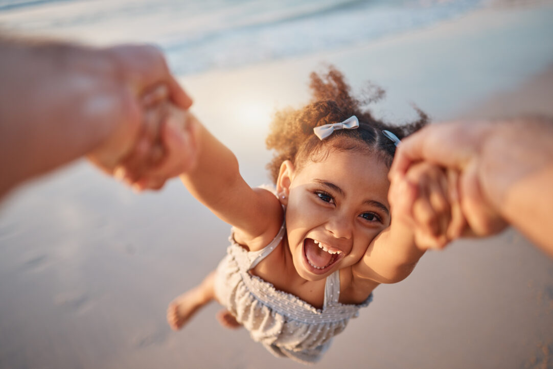 Girl child, spin and pov by ocean, portrait and smile for game, holding hands or speed in summer. Young female kid, parent and swing in air, sand or happy for family bonding, love or care in sunshine.